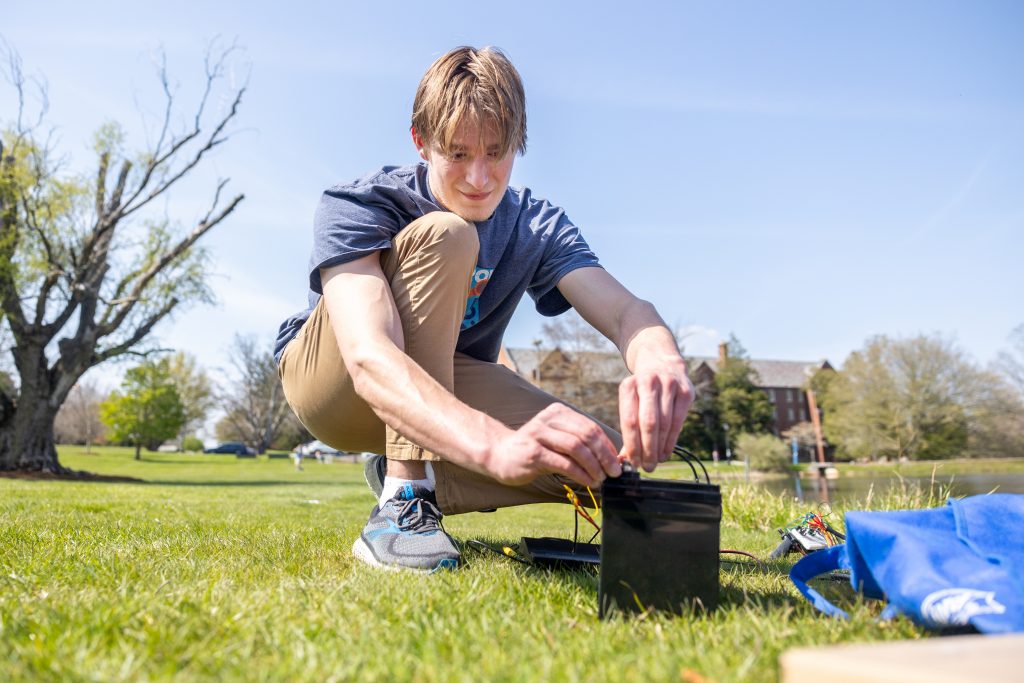 Oliver Mackinnon '25 plugs the wiring for the small underwater robot prototype built by UConn's Underwater Robotics Club into the car battery that powers it during a test run at Mirror Lake on April 28, 2024. (Sydney Herdle/UConn Photo)